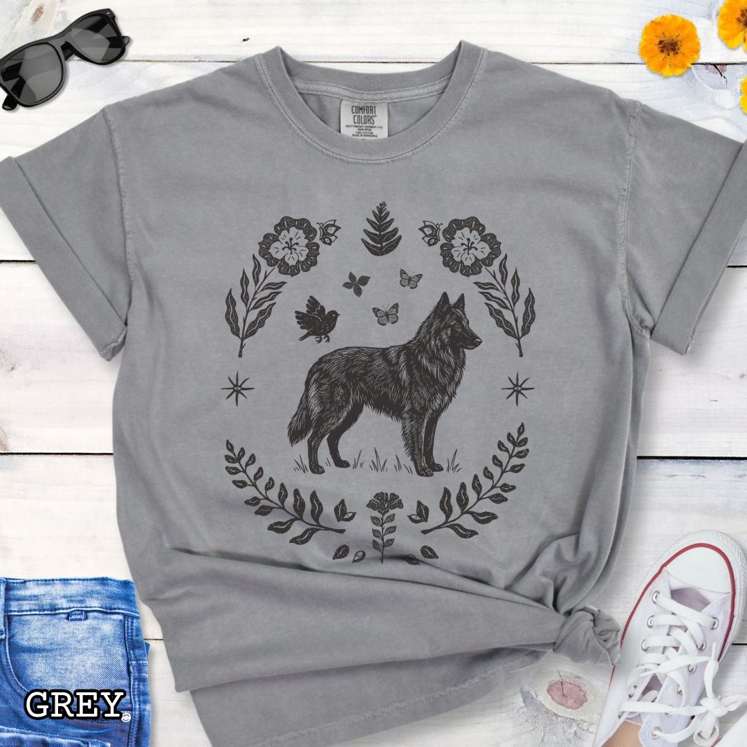 Grey Comfort Colors t-shirt for Belgian Sheepdog/Tervuren lovers, with folk art design of Belgian Shepherd surrounded by botanical elements, done in linocut monochrome style. Shown on a wooden surface accompanied by jeans, shoes and accessories.