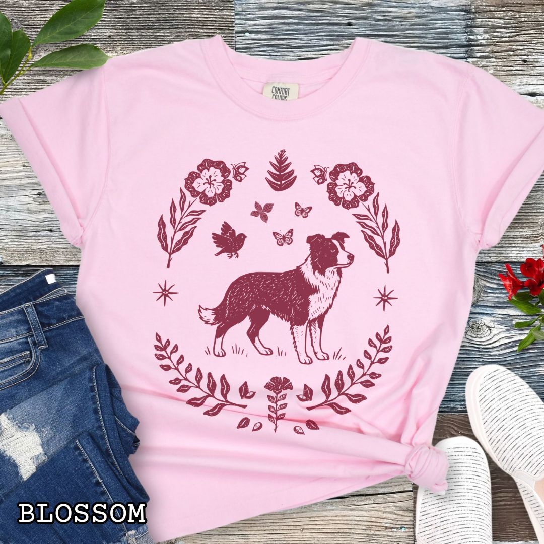 Pink t-shirt for Border Collie lovers with monochrome linocut-style design of a Border Collie dog surrounded by floral botanical elements, shown on a wooden surface with jeans and sneakers.