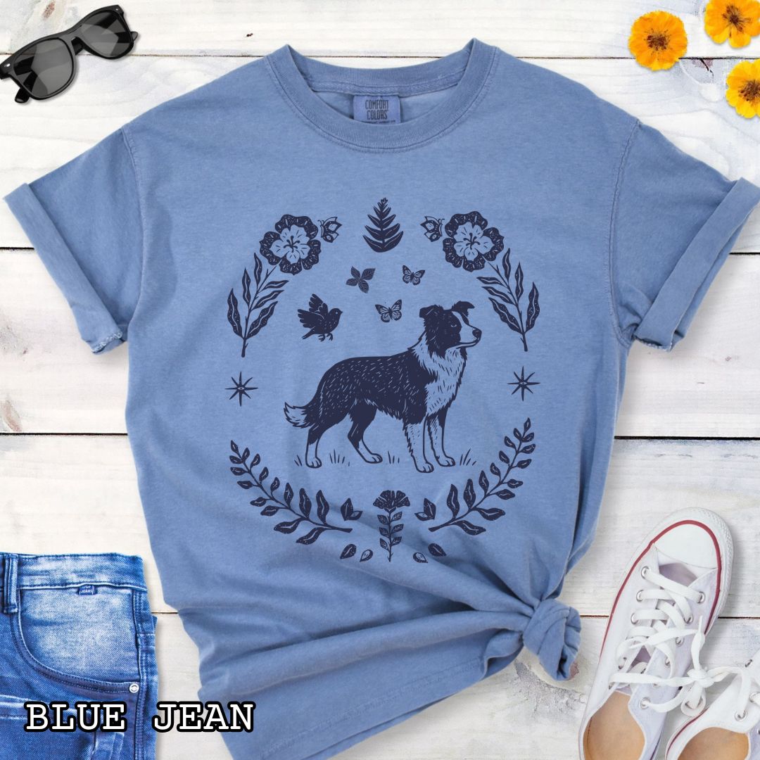 Blue t-shirt for Border Collie lovers with monochrome linocut-style design of a Border Collie dog surrounded by floral botanical elements, shown on a wooden surface with jeans and sneakers.