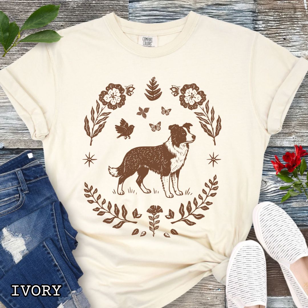Beige t-shirt for Border Collie lovers with monochrome linocut-style design of a Border Collie dog surrounded by floral botanical elements, shown on a wooden surface with jeans and sneakers.