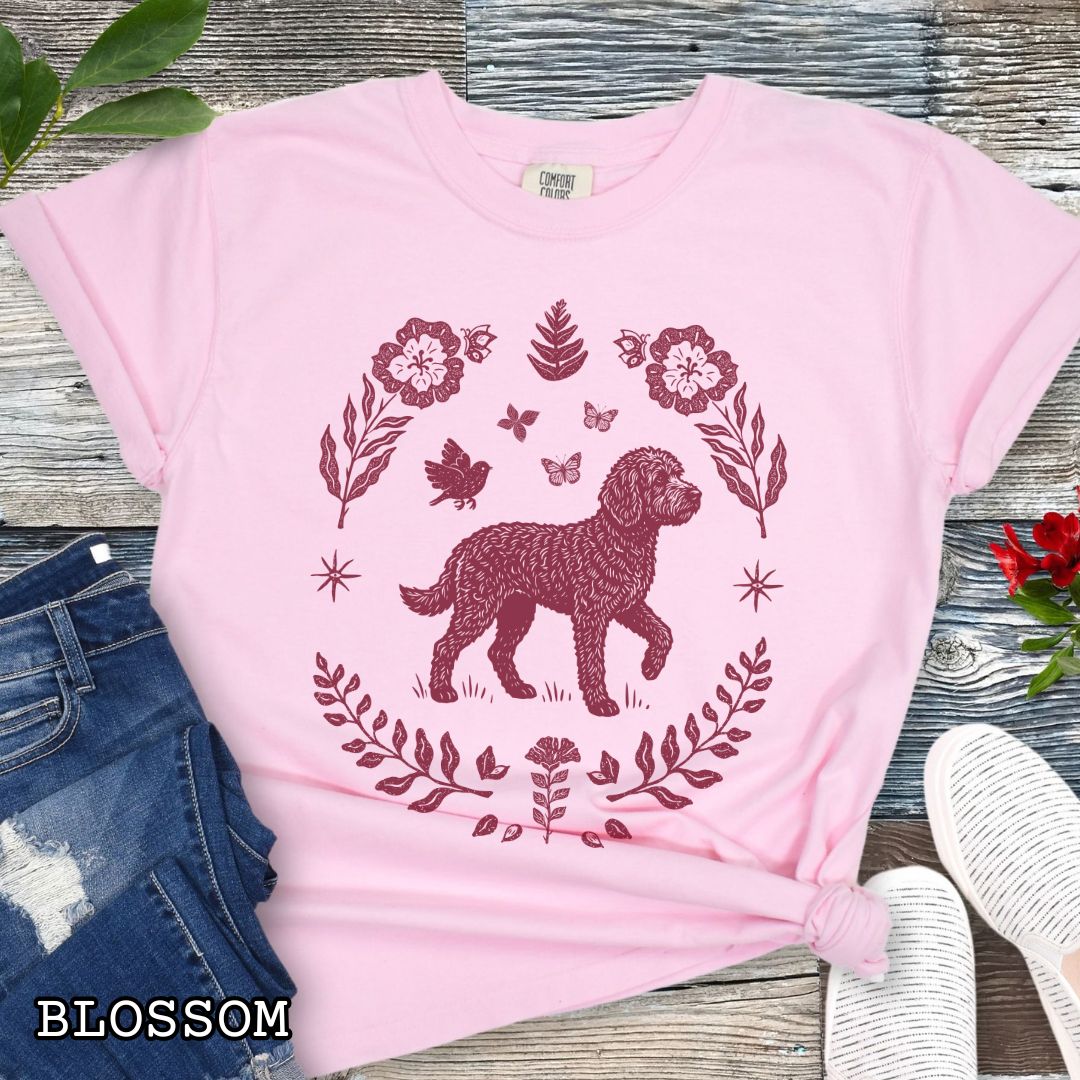 Pink Comfort Colors t-shirt for Doodle dog lovers, with folk art design of Goldendoodle surrounded by botanical elements, done in linocut monochrome style. Shown on a wooden surface accompanied by jeans, shoes and accessories.