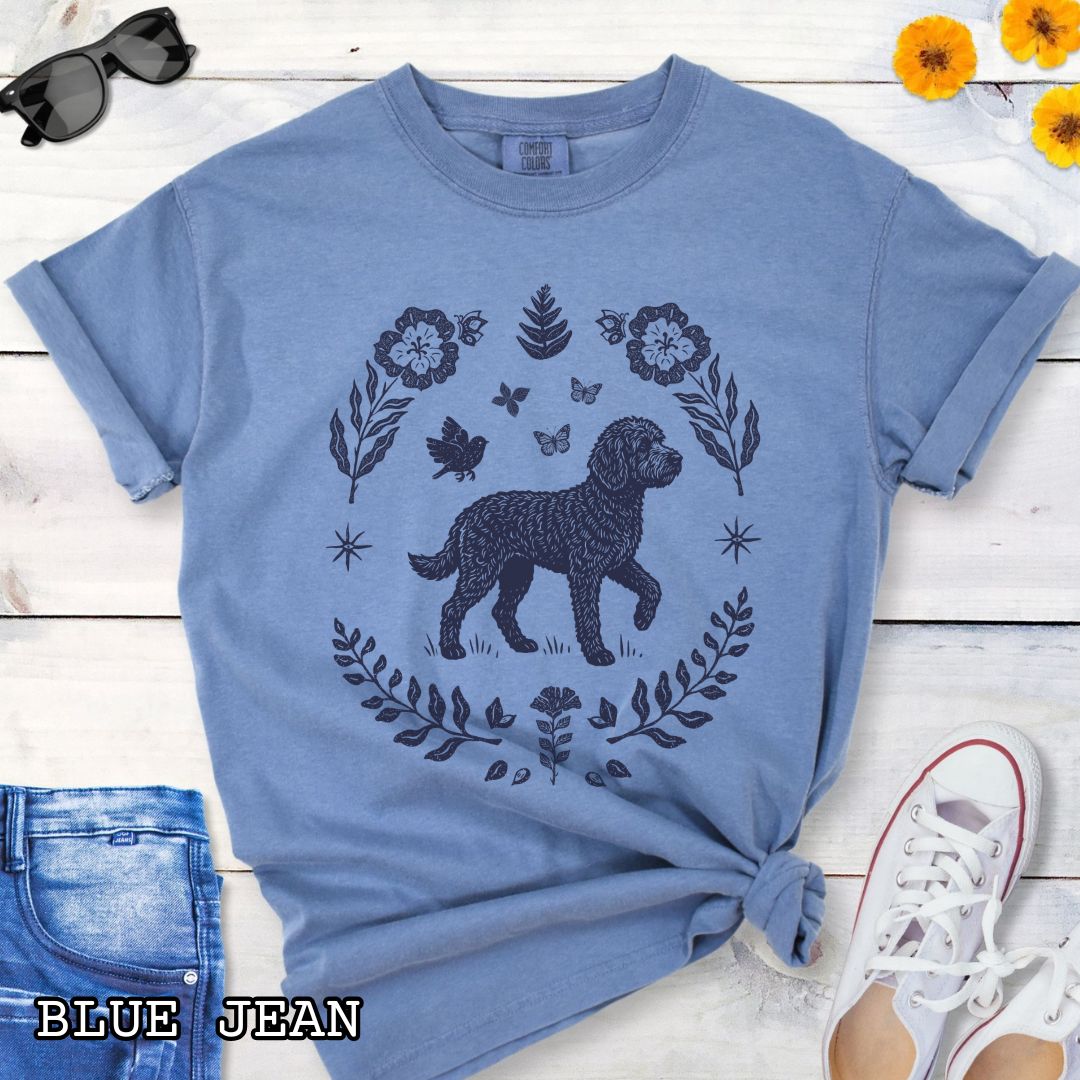 Blue Comfort Colors t-shirt for Doodle dog lovers, with folk art design of Bernedoodle surrounded by botanical elements, done in linocut monochrome style. Shown on a wooden surface accompanied by jeans, shoes and accessories.