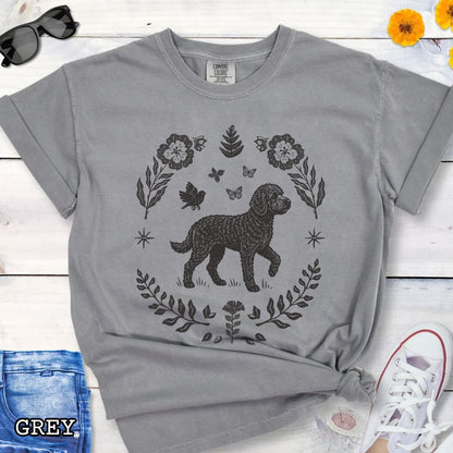 Grey Comfort Colors t-shirt for Doodle dog lovers, with folk art design of Doodle dog surrounded by botanical elements, done in linocut monochrome style. Shown on a wooden surface accompanied by jeans, shoes and accessories.