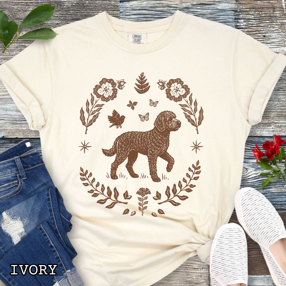 Beige Comfort Colors t-shirt for Doodle dog lovers, with folk art design of Labradoodle surrounded by botanical elements, done in linocut monochrome style. Shown on a wooden surface accompanied by jeans, shoes and accessories.