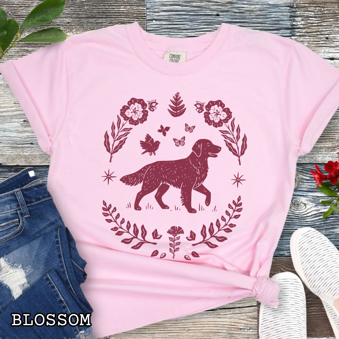 Pink Comfort Colors t-shirt for Golden Retriever lovers with linocut-style monochrome design of an Golden dog surrounded by floral botanical elements, shown on a wooden surface with jeans and flowers.