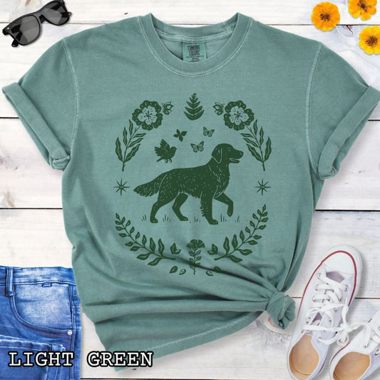 Green Comfort Colors t-shirt for Golden Retriever lovers with linocut-style monochrome design of an Golden dog surrounded by floral botanical elements, shown on a wooden surface with jeans and flowers.