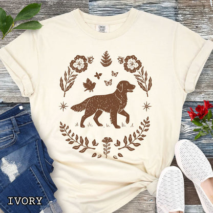 Ivory Comfort Colors t-shirt for Golden Retriever lovers with linocut-style monochrome design of an Golden dog surrounded by floral botanical elements, shown on a wooden surface with jeans and flowers.