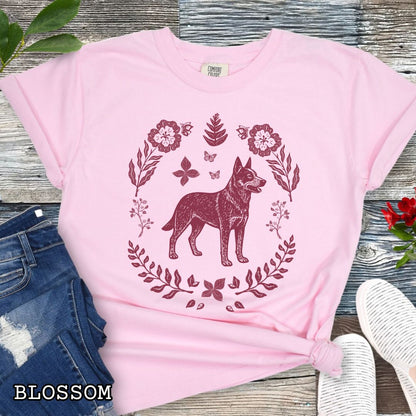 Pink Comfort Colors t-shirt for Australian Cattle Dog lovers with monochrome linocut-style design of a Heeler dog surrounded by floral botanical elements, shown on a wooden surface with jeans and sneakers.