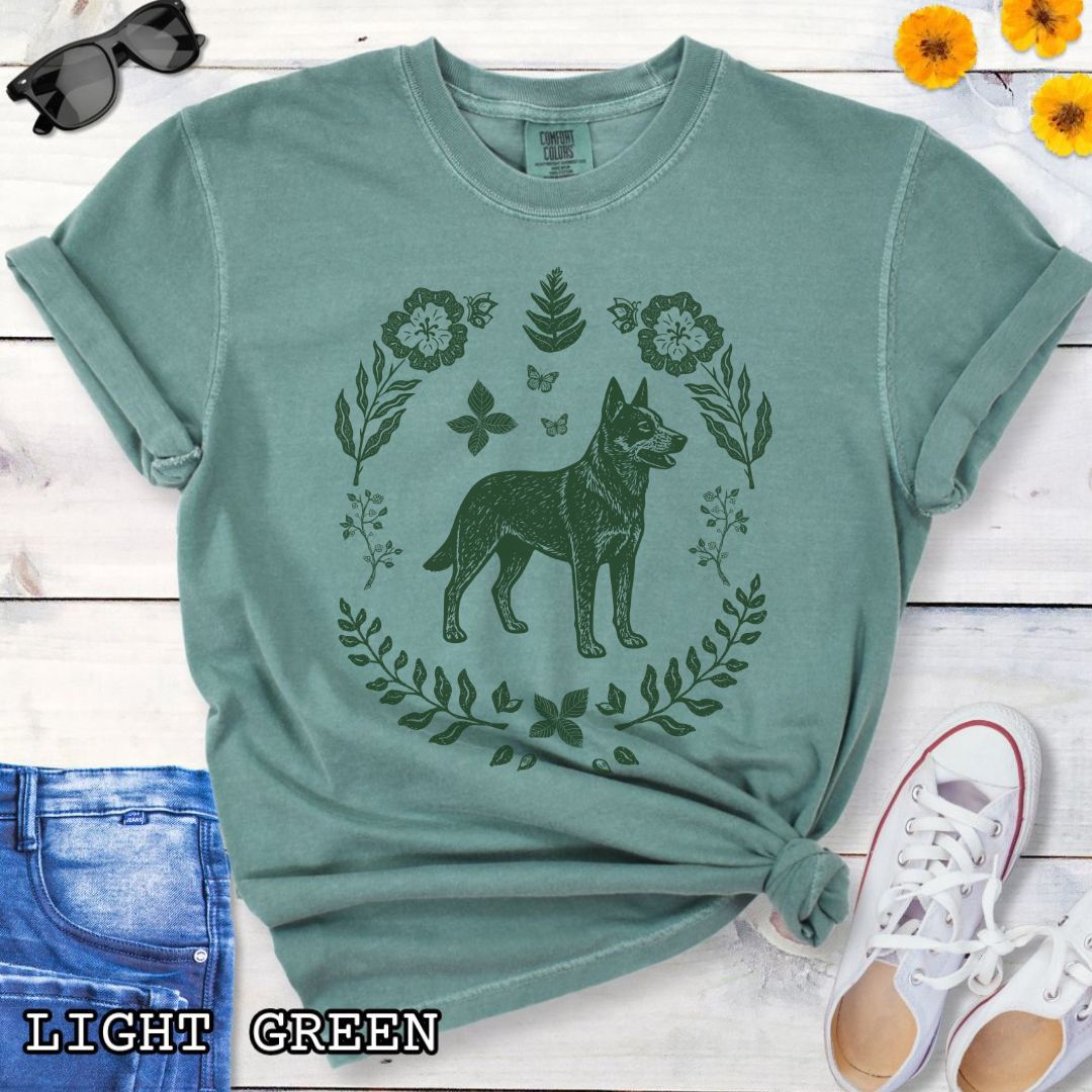 Green Comfort Colors t-shirt for Australian Cattle Dog lovers with monochrome linocut-style design of a Heeler dog surrounded by floral botanical elements, shown on a wooden surface with jeans and sneakers.