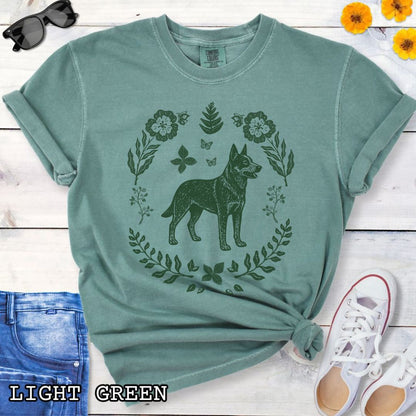 Green Comfort Colors t-shirt for Australian Cattle Dog lovers with monochrome linocut-style design of a Heeler dog surrounded by floral botanical elements, shown on a wooden surface with jeans and sneakers.