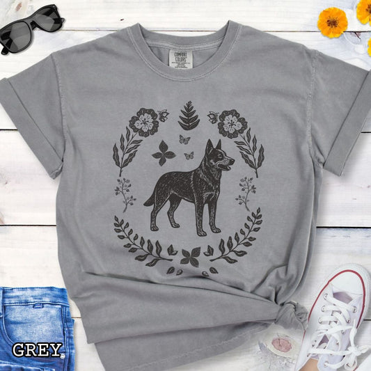 Grey Comfort Colors t-shirt for Australian Cattle Dog lovers with monochrome linocut-style design of a Heeler dog surrounded by floral botanical elements, shown on a wooden surface with jeans and sneakers.