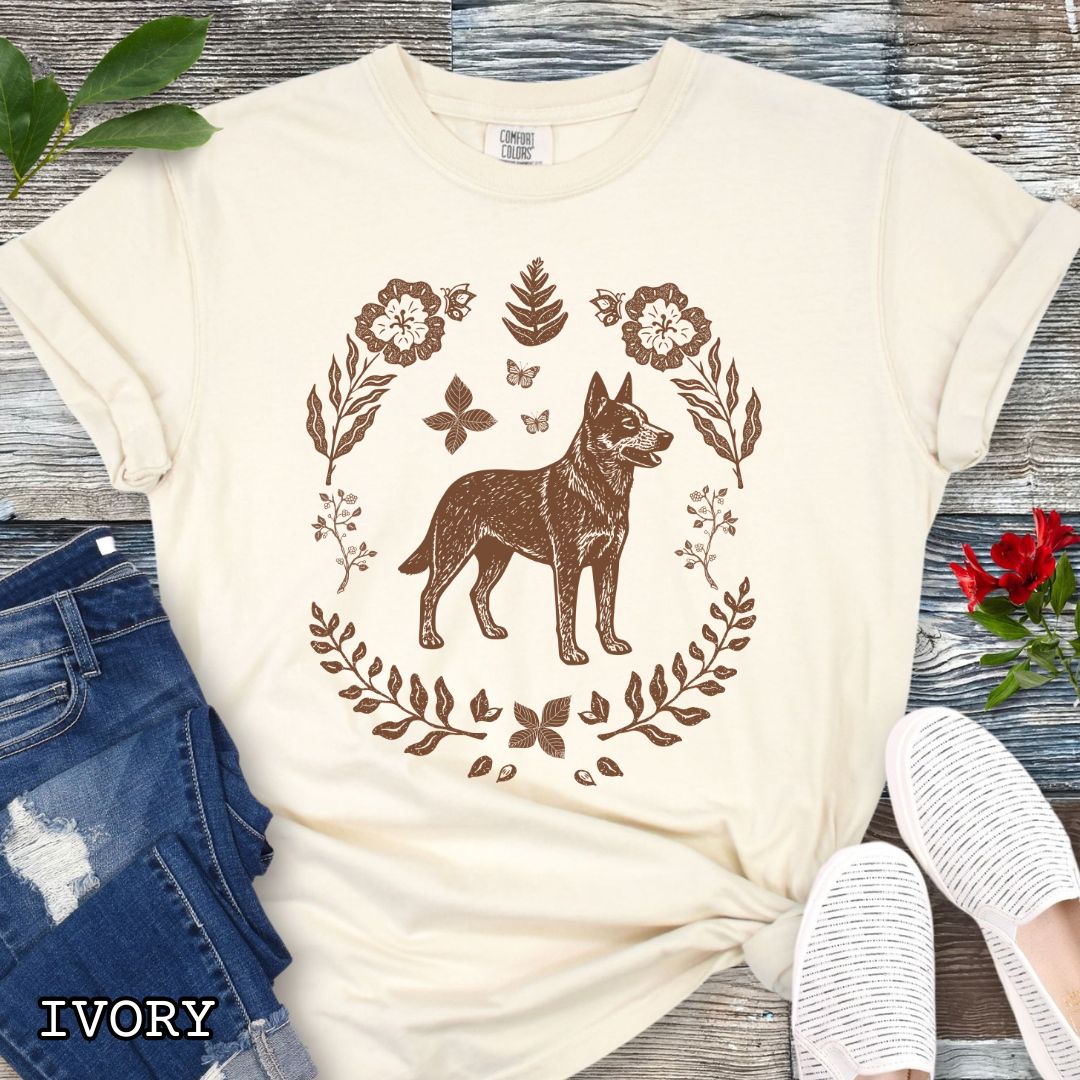Beige Comfort Colors t-shirt for Australian Cattle Dog lovers with monochrome linocut-style design of a Heeler dog surrounded by floral botanical elements, shown on a wooden surface with jeans and sneakers.