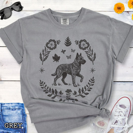 Grey Comfort Colors t-shirt for Belgian Laekenois lovers, with folk art design of Belgian Laekenois dog surrounded by botanical elements, done in linocut monochrome style. Shown on a wooden surface accompanied by jeans, shoes and accessories.