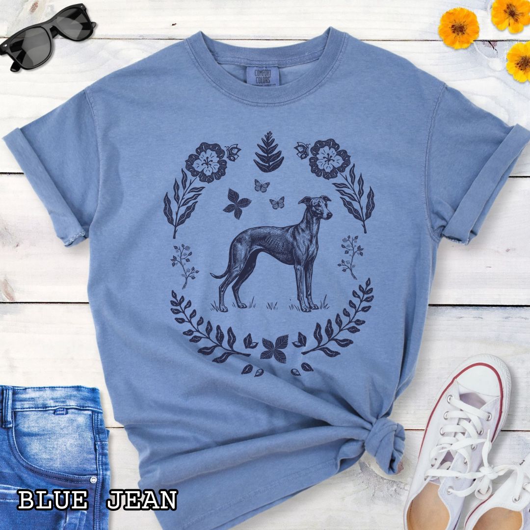 Blue Comfort Colors t-shirt for sighthound lovers with monochrome linocut-style design of a Whippet/Italian Greyhound dog surrounded by floral botanical elements, shown on a wooden surface with jeans and sneakers.