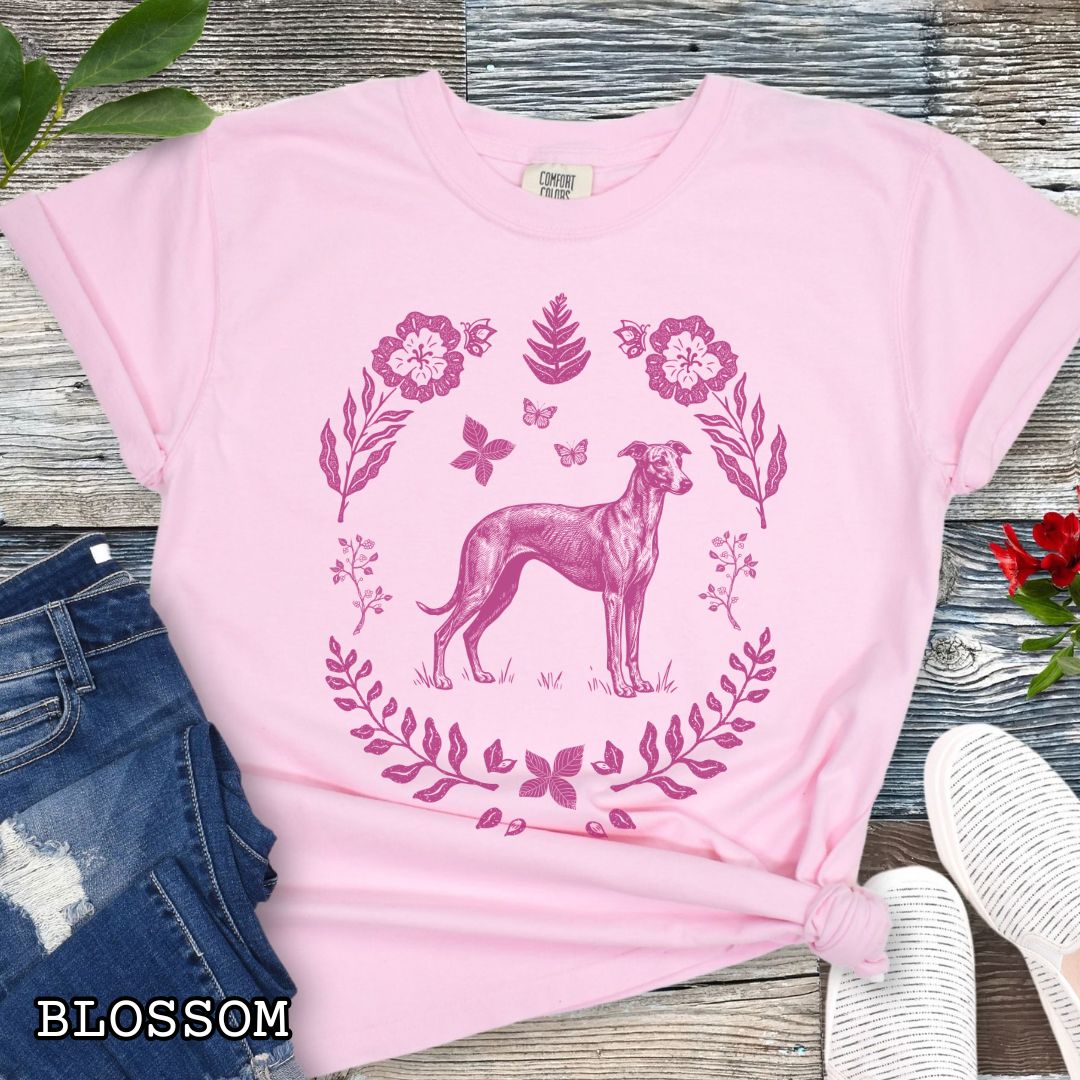Pink Comfort Colors t-shirt for sighthound lovers with monochrome linocut-style design of a Whippet/Italian Greyhound dog surrounded by floral botanical elements, shown on a wooden surface with jeans and sneakers.