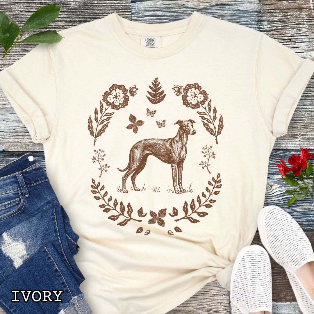 Beige Comfort Colors t-shirt for sighthound lovers with monochrome linocut-style design of a Whippet/Italian Greyhound dog surrounded by floral botanical elements, shown on a wooden surface with jeans and sneakers.