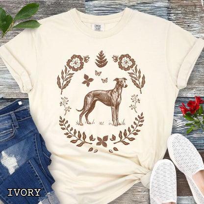 Beige Comfort Colors t-shirt for sighthound lovers with monochrome linocut-style design of a Whippet/Italian Greyhound dog surrounded by floral botanical elements, shown on a wooden surface with jeans and sneakers.