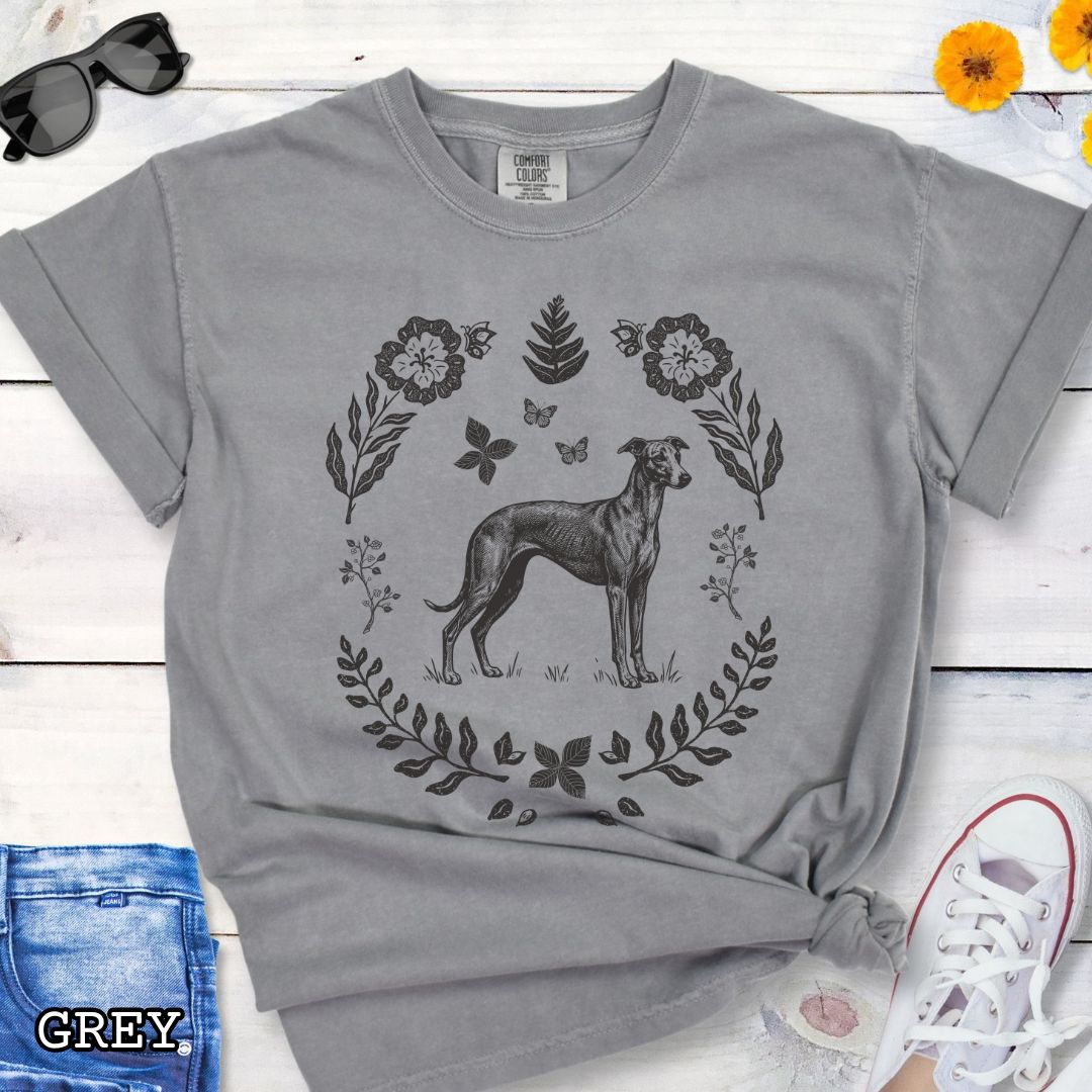 Grey Comfort Colors t-shirt for sighthound lovers with monochrome linocut-style design of a Whippet/Italian Greyhound dog surrounded by floral botanical elements, shown on a wooden surface with jeans and sneakers.