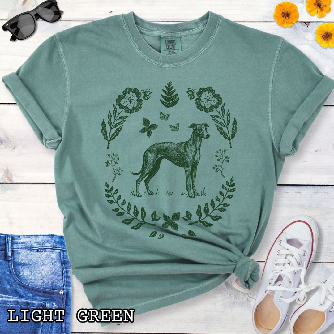 Green Comfort Colors t-shirt for sighthound lovers with monochrome linocut-style design of a Whippet/Italian Greyhound dog surrounded by floral botanical elements, shown on a wooden surface with jeans and sneakers.