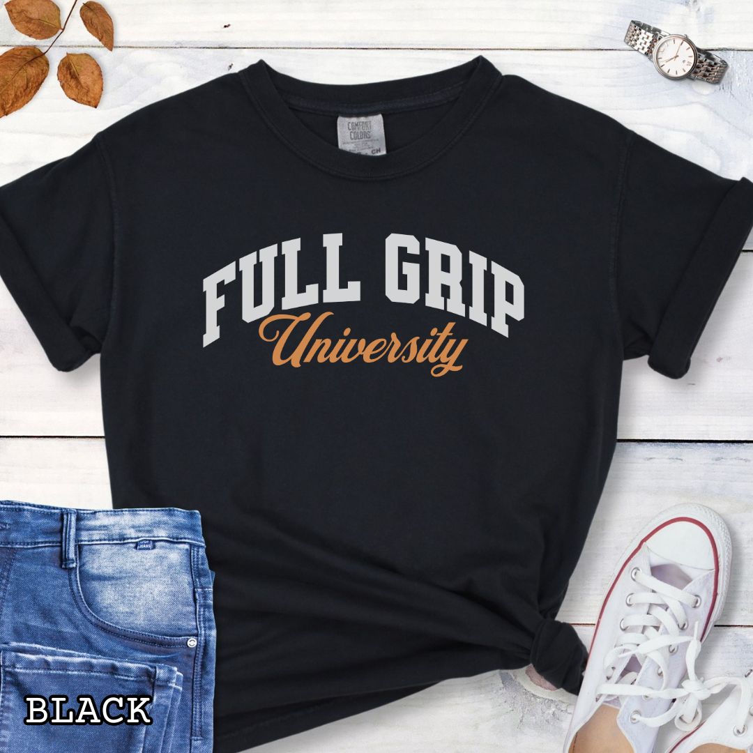Black Comfort Colors t-shirt for bitework dog trainers with 'FULL GRIP University' text in college style lettering, shown on a wooden surface with jeans and sneakers.