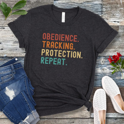 Dark grey protection dog sports t-shirt with colorful text 'OBEDIENCE. TRACKING. PROTECTION. REPEAT.', presented on a wooden surface with jeans and shoes.
