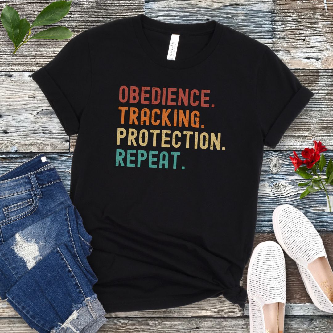 Black protection dog sports t-shirt with colorful text 'OBEDIENCE. TRACKING. PROTECTION. REPEAT.', presented on a wooden surface with jeans and shoes.