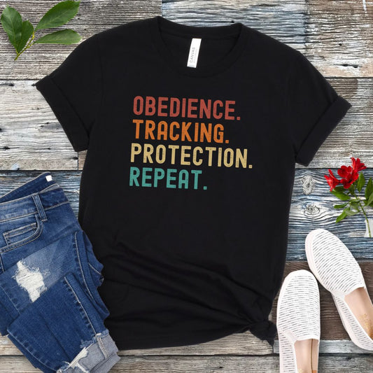 Black protection dog sports t-shirt with colorful text 'OBEDIENCE. TRACKING. PROTECTION. REPEAT.', presented on a wooden surface with jeans and shoes.