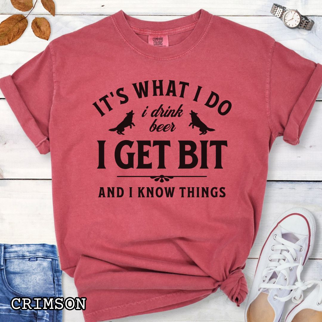 Red Comfort Colors K9 helper t-shirt with bitework graphic and the humorous text 'It's What I Do, I drink beer, I get bit, and I know things', shown on a wooden surface with shoes and jeans.