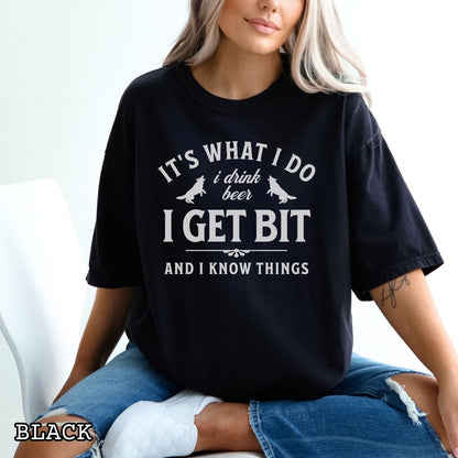 Black Comfort Colors K9 helper t-shirt with bitework graphic and the humorous text 'It's What I Do, I drink beer, I get bit, and I know things', shown on a wooden surface with shoes and jeans.