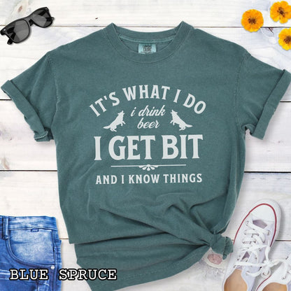 Green Comfort Colors K9 helper t-shirt with bitework graphic and the humorous text 'It's What I Do, I drink beer, I get bit, and I know things', shown on a wooden surface with shoes and jeans.