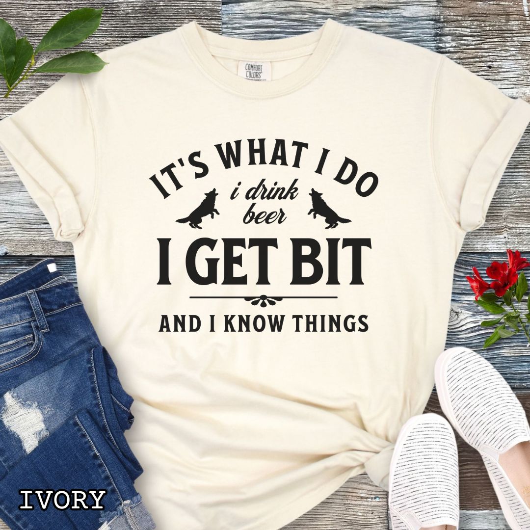Ivory Comfort Colors K9 helper t-shirt with bitework graphic and the humorous text 'It's What I Do, I drink beer, I get bit, and I know things', shown on a wooden surface with shoes and jeans.