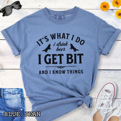 Blue Comfort Colors K9 helper t-shirt with bitework graphic and the humorous text 'It's What I Do, I drink beer, I get bit, and I know things', shown on a wooden surface with shoes and jeans.