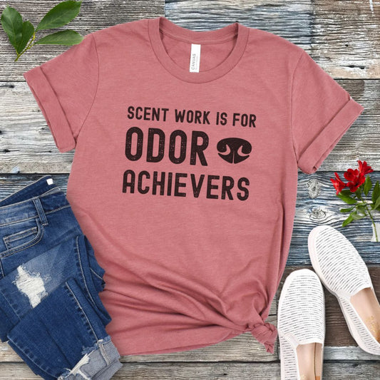 Mauve canine scent work t-shirt with 'Scent work is for odor achievers' text on a wooden surface with jeans and shoes.