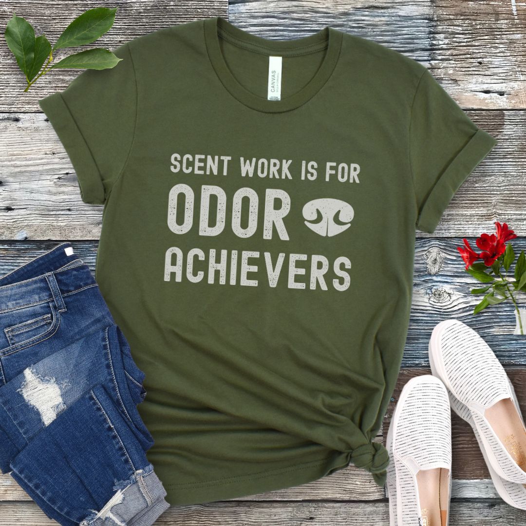 Military green canine scent work t-shirt with 'Scent work is for odor achievers' text on a wooden surface with jeans and shoes.
