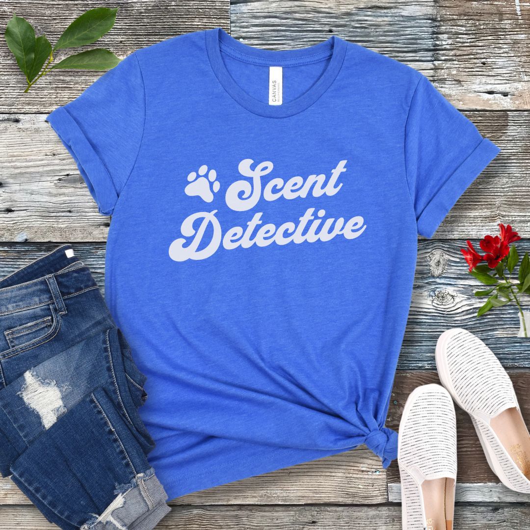 Royal blue K9 nosework t-shirt with 'Scent Detective' text design in retro font, presented on a wooden surface with jeans and shoes.
