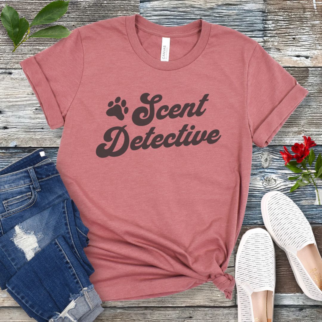 Mauve canine scent work t-shirt with 'Scent Detective' text design in retro font, presented on a wooden surface with jeans and shoes.