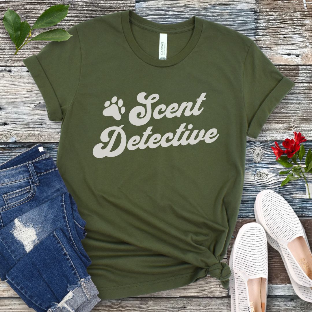 Military green dog nosework t-shirt with 'Scent Detective' text design in retro font, presented on a wooden surface with jeans and shoes.