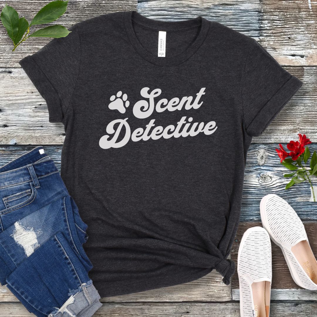 Dark grey detection dog training t-shirt with 'Scent Detective' text design in retro font, presented on a wooden surface with jeans and shoes.
