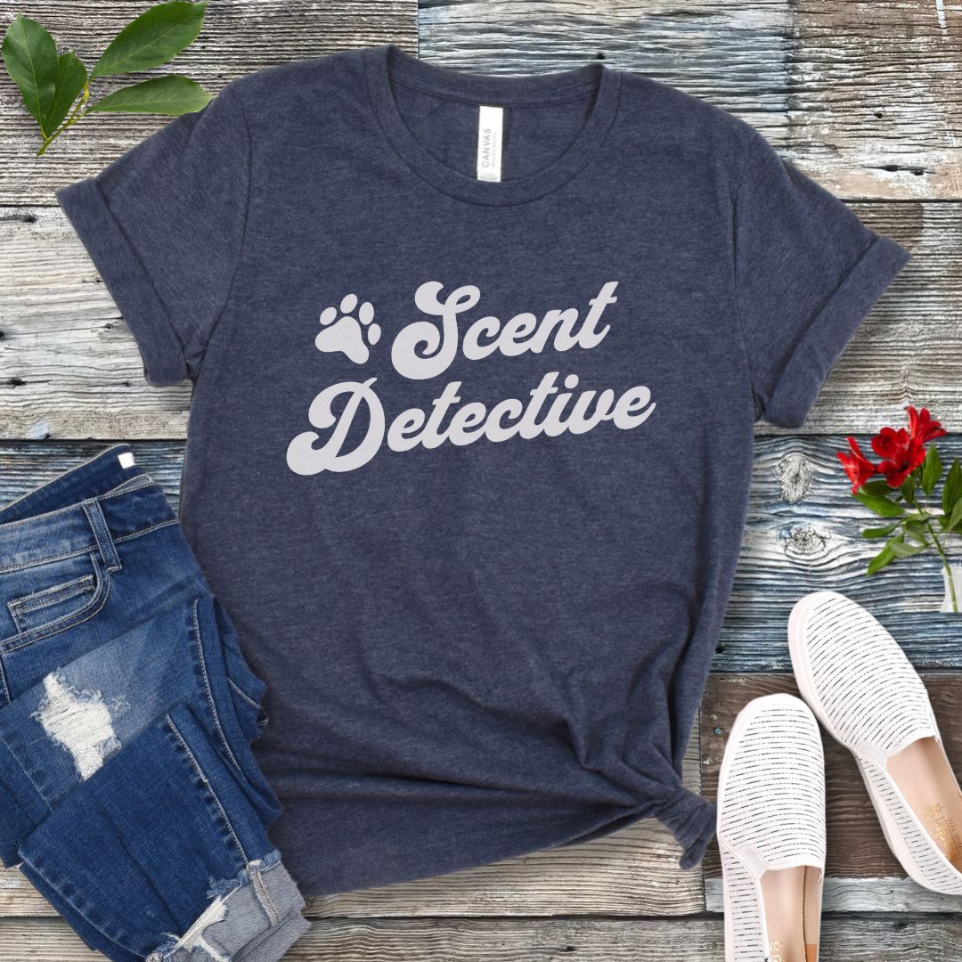 Heather navy dog nosework t-shirt with 'Scent Detective' text design in retro font, presented on a wooden surface with jeans and shoes.