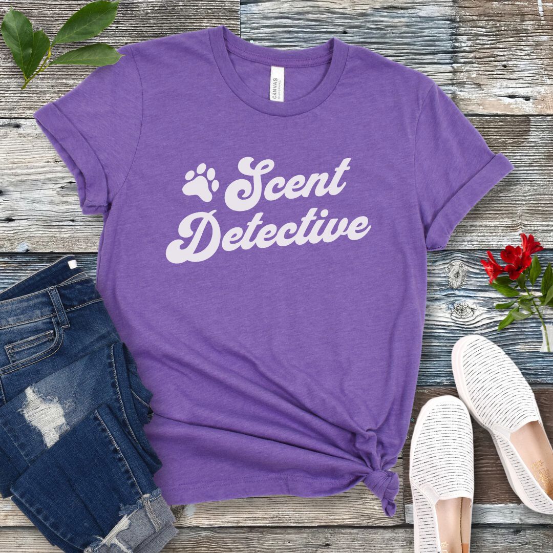 Purple K9 nosework t-shirt with 'Scent Detective' text design in retro font, presented on a wooden surface with jeans and shoes.