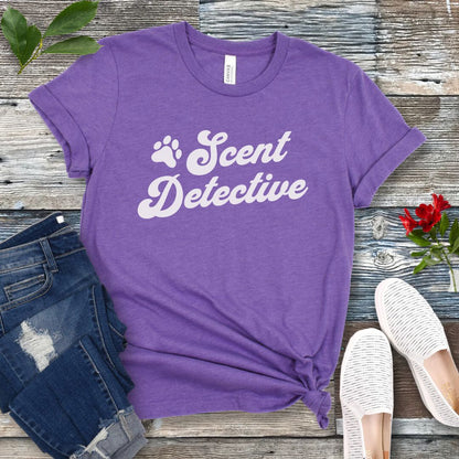 Purple K9 nosework t-shirt with 'Scent Detective' text design in retro font, presented on a wooden surface with jeans and shoes.