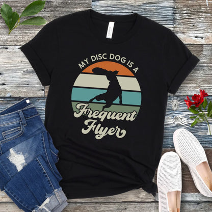 Black unisex disc dog t-shirt with colorful vintage sunset, leaping dog catching a disc, and the humorous text 'My Disc dog is a frequent flyer'. 