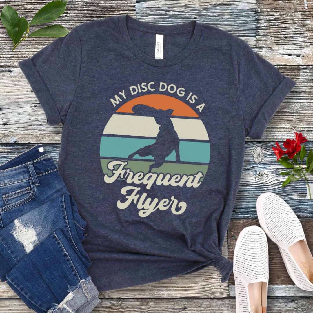 Heather navy unisex disc dog t-shirt with colorful vintage sunset, leaping dog catching a disc, and the humorous text 'My Disc dog is a frequent flyer'. 