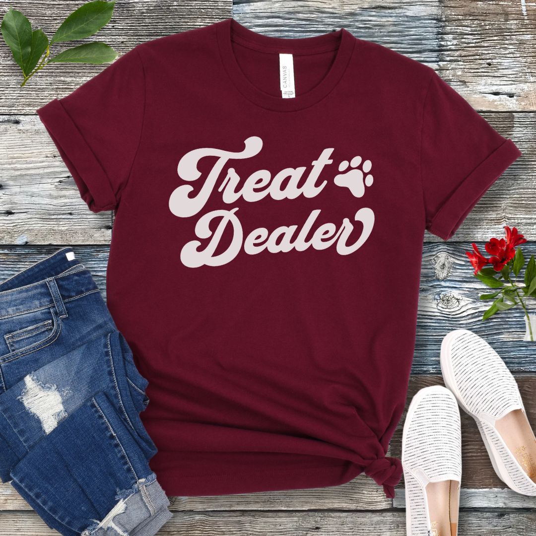 A maroon T-shirt with the retro font with the text 'Treat Dealer' printed on the front, accompanied by a paw print. The shirt is displayed on a wooden background with a pair of jeans and white shoes.