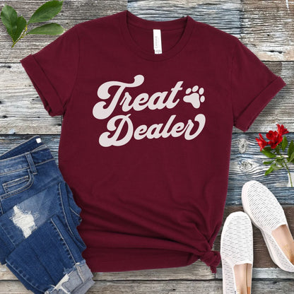 A maroon T-shirt with the retro font with the text 'Treat Dealer' printed on the front, accompanied by a paw print. The shirt is displayed on a wooden background with a pair of jeans and white shoes.