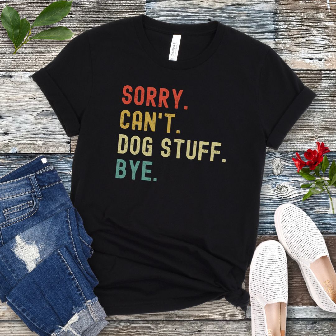 A black unisex short sleeve T-shirt with colorful stacked text 'SORRY. CAN'T. DOG STUFF. BYE.', presented on a wooden surface with jeans and shoes.