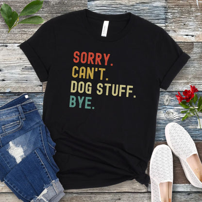 A black unisex short sleeve T-shirt with colorful stacked text 'SORRY. CAN'T. DOG STUFF. BYE.', presented on a wooden surface with jeans and shoes.