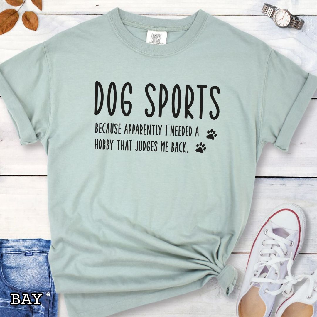 Light green Comfort Colors t-shirt with humorous text 'Dog Sports, Because Apparently I Needed a Hobby That Judges Me Back' accompanied by paw prints, shown on a wooden surface with jeans and flowers.