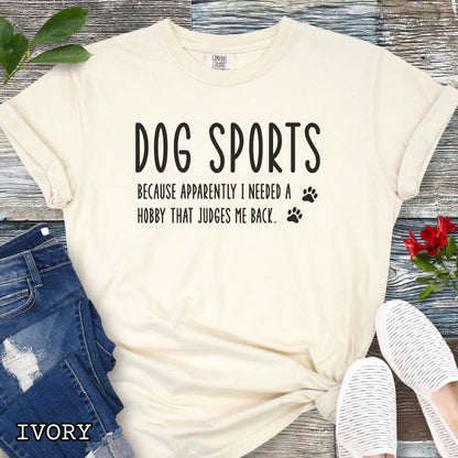Beige Comfort Colors t-shirt with humorous text 'Dog Sports, Because Apparently I Needed a Hobby That Judges Me Back' accompanied by paw prints, shown on a wooden surface with jeans and flowers.