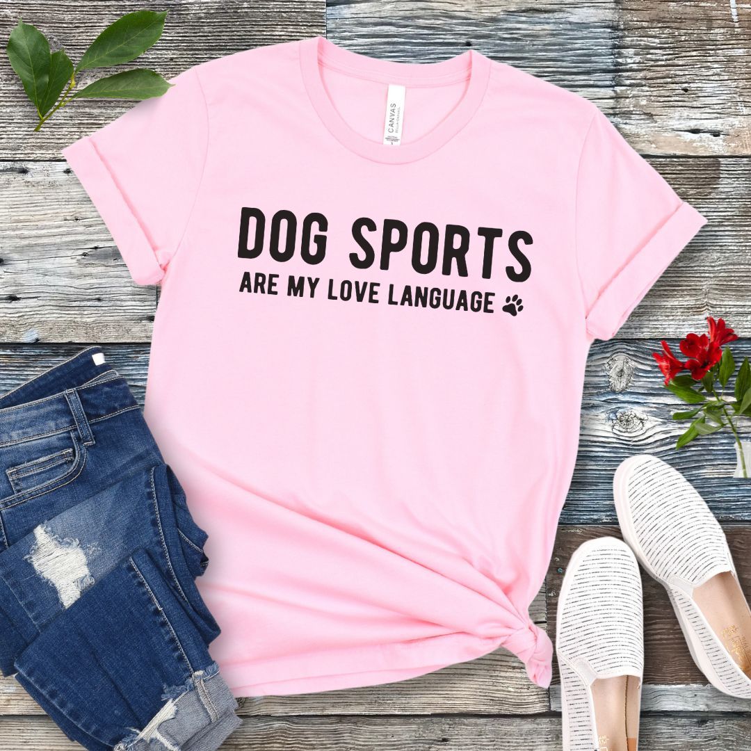 Pink t-shirt with 'Dog Sports are my love language' text on a wooden surface with jeans and shoes.